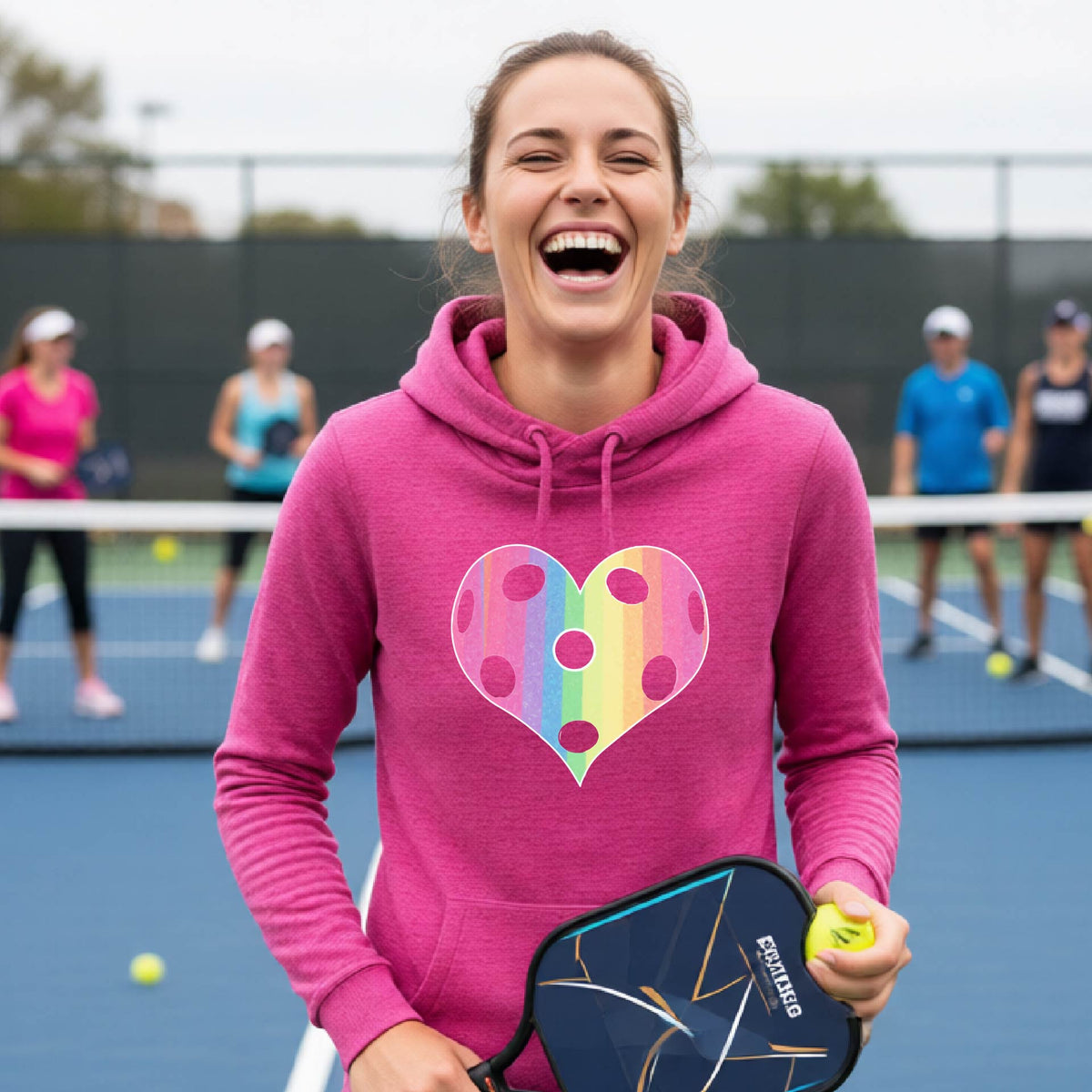 A woman laughing on a pickleball court, wearing a pink cowl-neck hoodie pullover with a graphic of a brightly rainbow-colored heart designed like a pickleball.