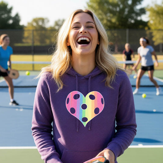A woman laughing on a pickleball court, wearing a purple hoodie pullover with a graphic of a brightly rainbow-colored heart designed like a pickleball.
