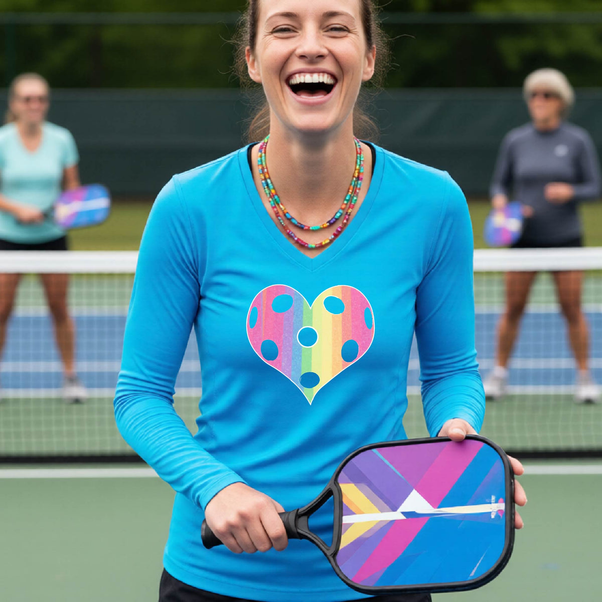 A woman laughing on a pickleball court, wearing a bright blue long-sleeve t-shirt with a graphic of a brightly rainbow-colored heart designed like a pickleball.