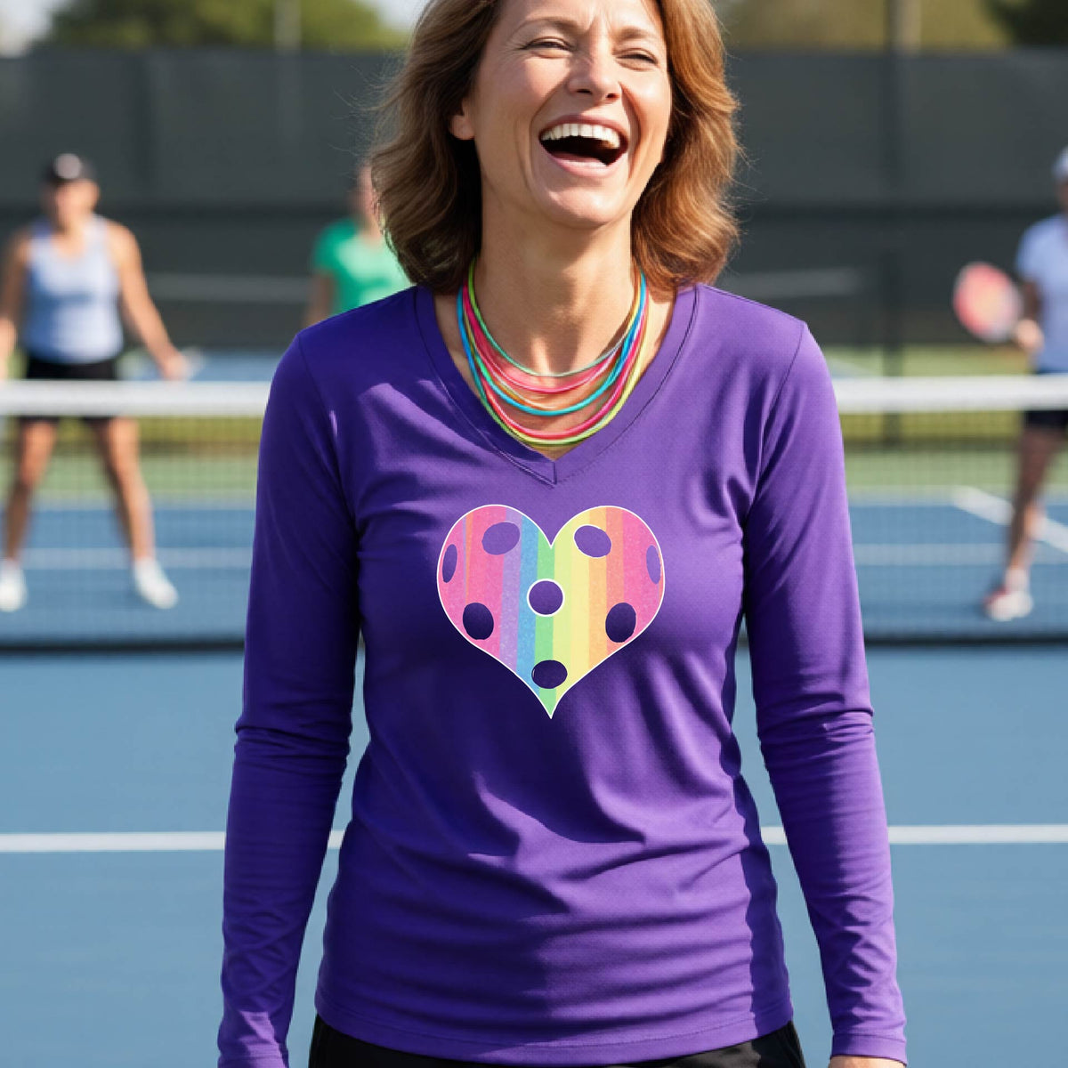 A woman laughing on a pickleball court, wearing a purple long-sleeve t-shirt with a graphic of a brightly rainbow-colored heart designed like a pickleball.