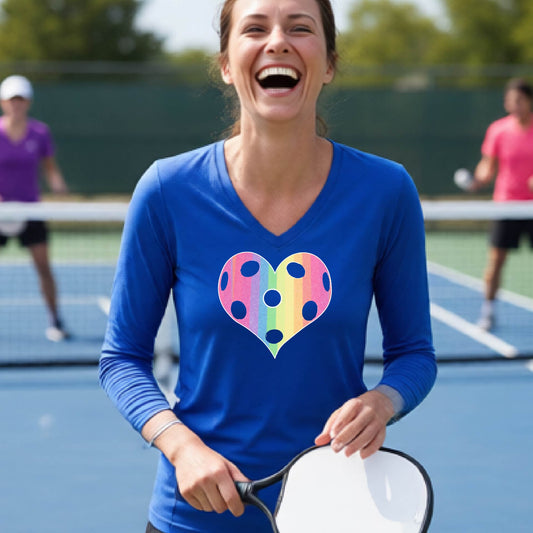 A woman laughing on a pickleball court, wearing a royal blue long-sleeve t-shirt with a graphic of a brightly rainbow-colored heart designed like a pickleball.