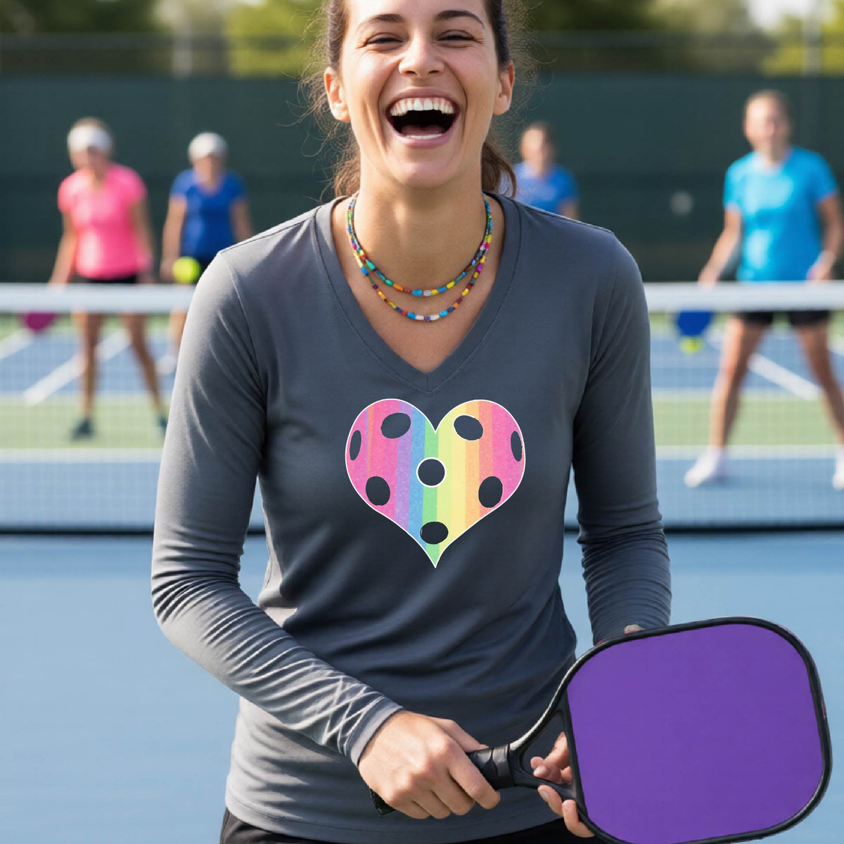 A woman laughing on a pickleball court, wearing a dark gray long-sleeve t-shirt with a graphic of a brightly rainbow-colored heart designed like a pickleball.