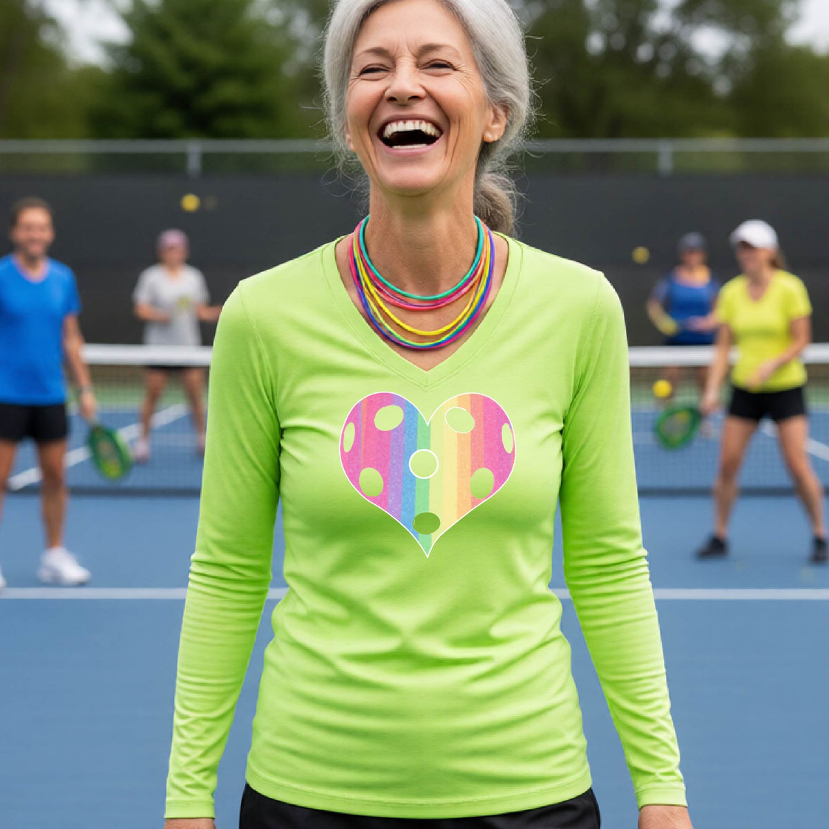 A woman laughing on a pickleball court, wearing a lime green long-sleeve t-shirt with a graphic of a brightly rainbow-colored heart designed like a pickleball.