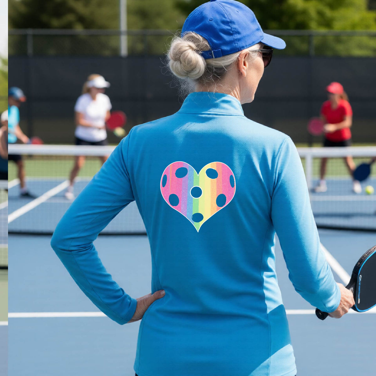 A woman on a pickleball court, facing backwards, wearing a bright blue quarter-zip athletic shirt with a graphic of a brightly rainbow-colored heart designed like a pickleball.