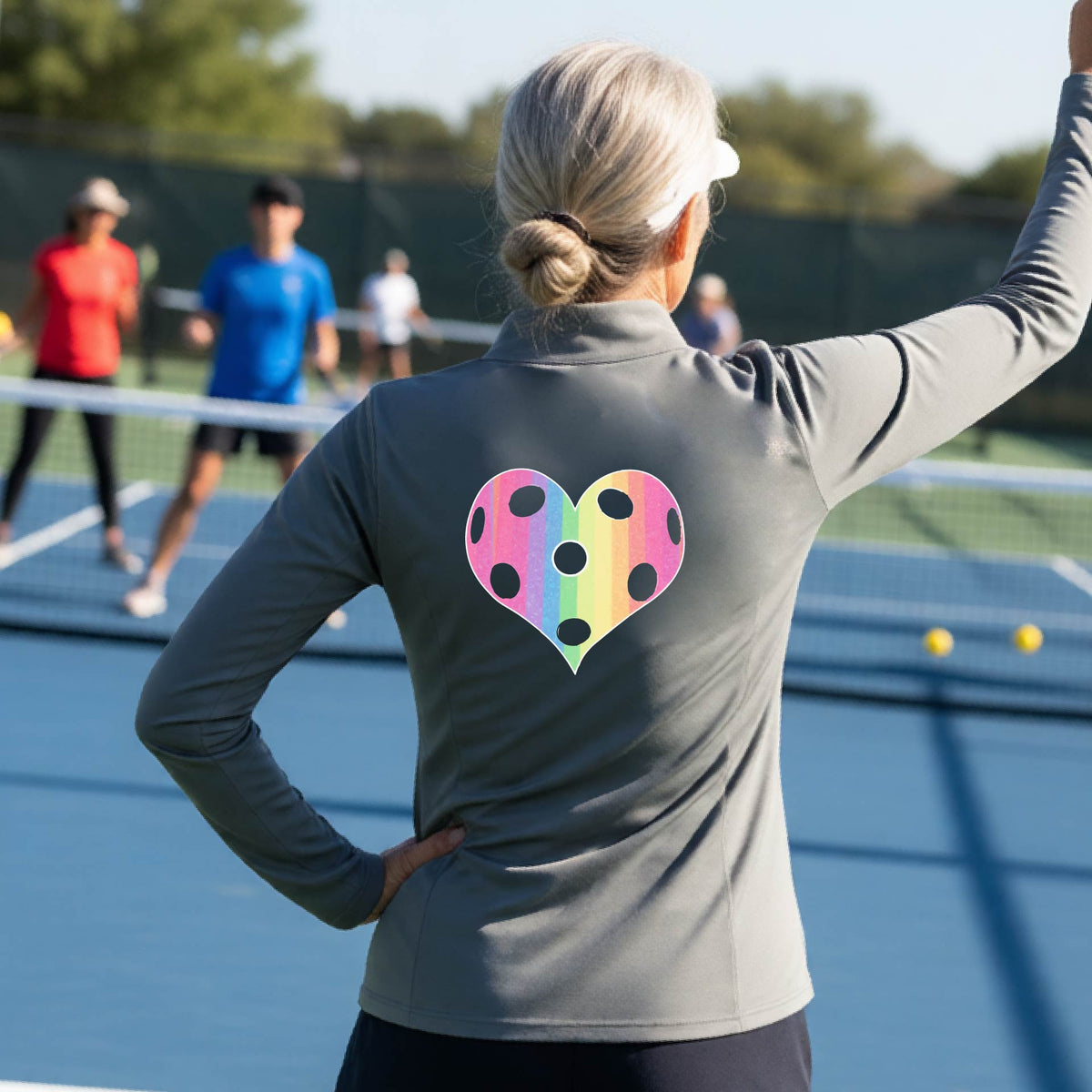 A woman on a pickleball court, facing backwards, wearing a gray quarter-zip athletic shirt with a graphic of a brightly rainbow-colored heart designed like a pickleball.