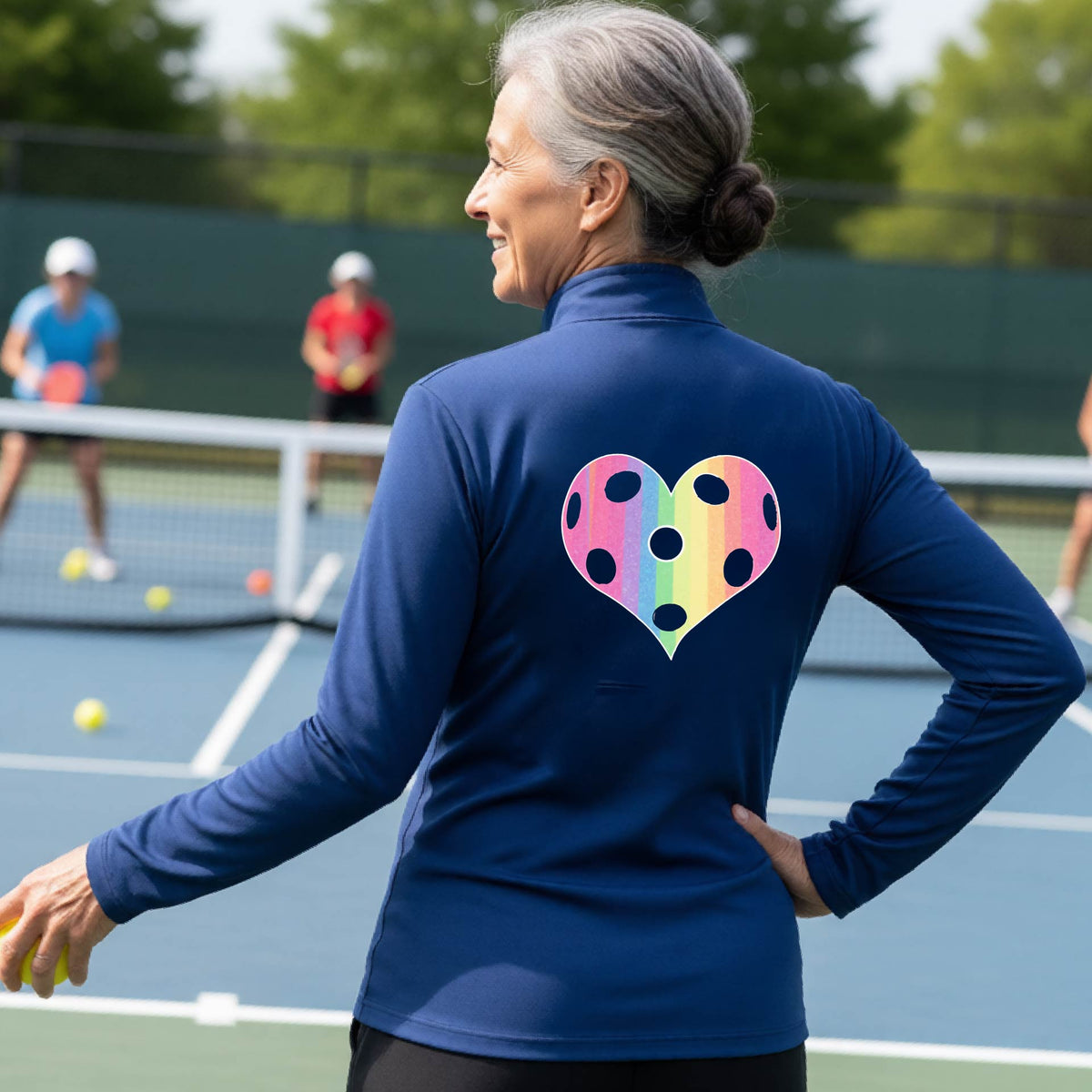 A woman on a pickleball court, facing backwards, wearing a royal blue quarter-zip athletic shirt with a graphic of a brightly rainbow-colored heart designed like a pickleball.
