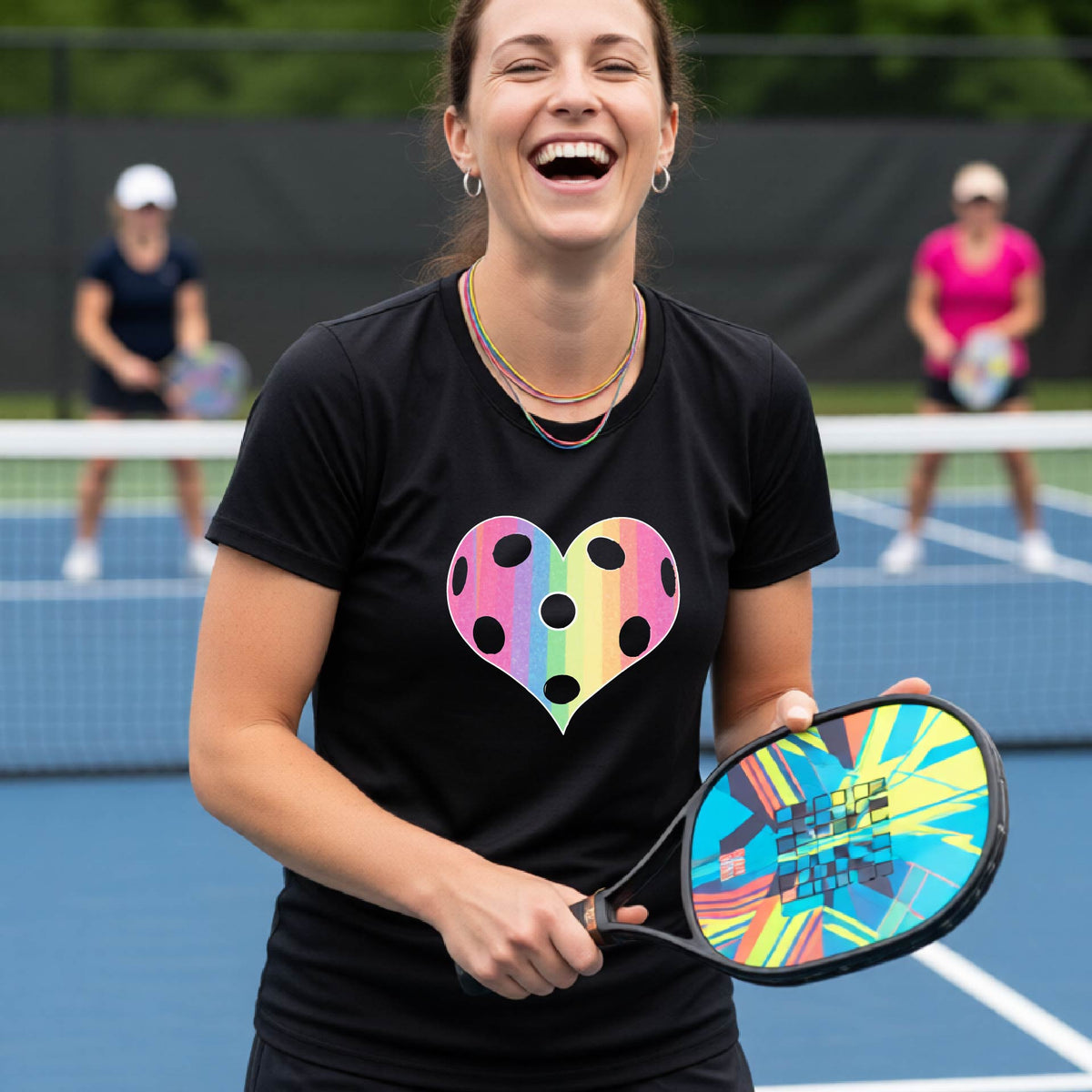 A woman laughing on a pickleball court, wearing a black t-shirt with a graphic of a brightly rainbow-colored heart designed like a pickleball.