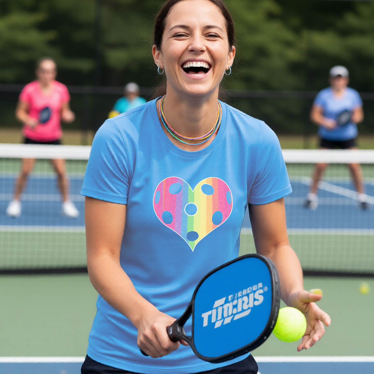 A woman laughing on a pickleball court, wearing a blue t-shirt with a graphic of a brightly rainbow-colored heart designed like a pickleball.