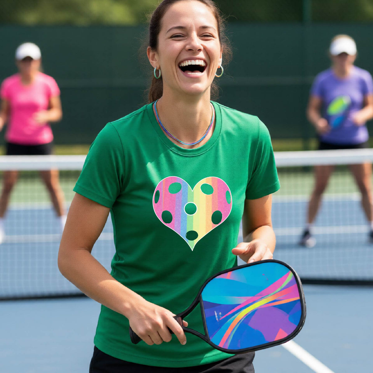 A woman laughing on a pickleball court, wearing a green t-shirt with a graphic of a brightly rainbow-colored heart designed like a pickleball.