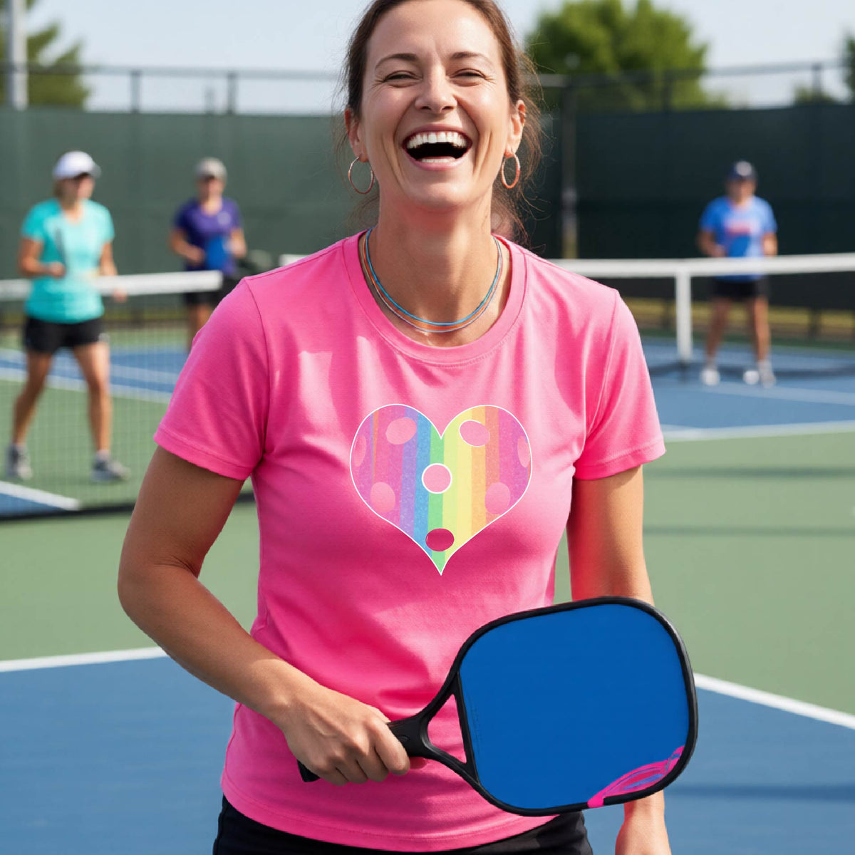 A woman laughing on a pickleball court, wearing a pink t-shirt with a graphic of a brightly rainbow-colored heart designed like a pickleball.