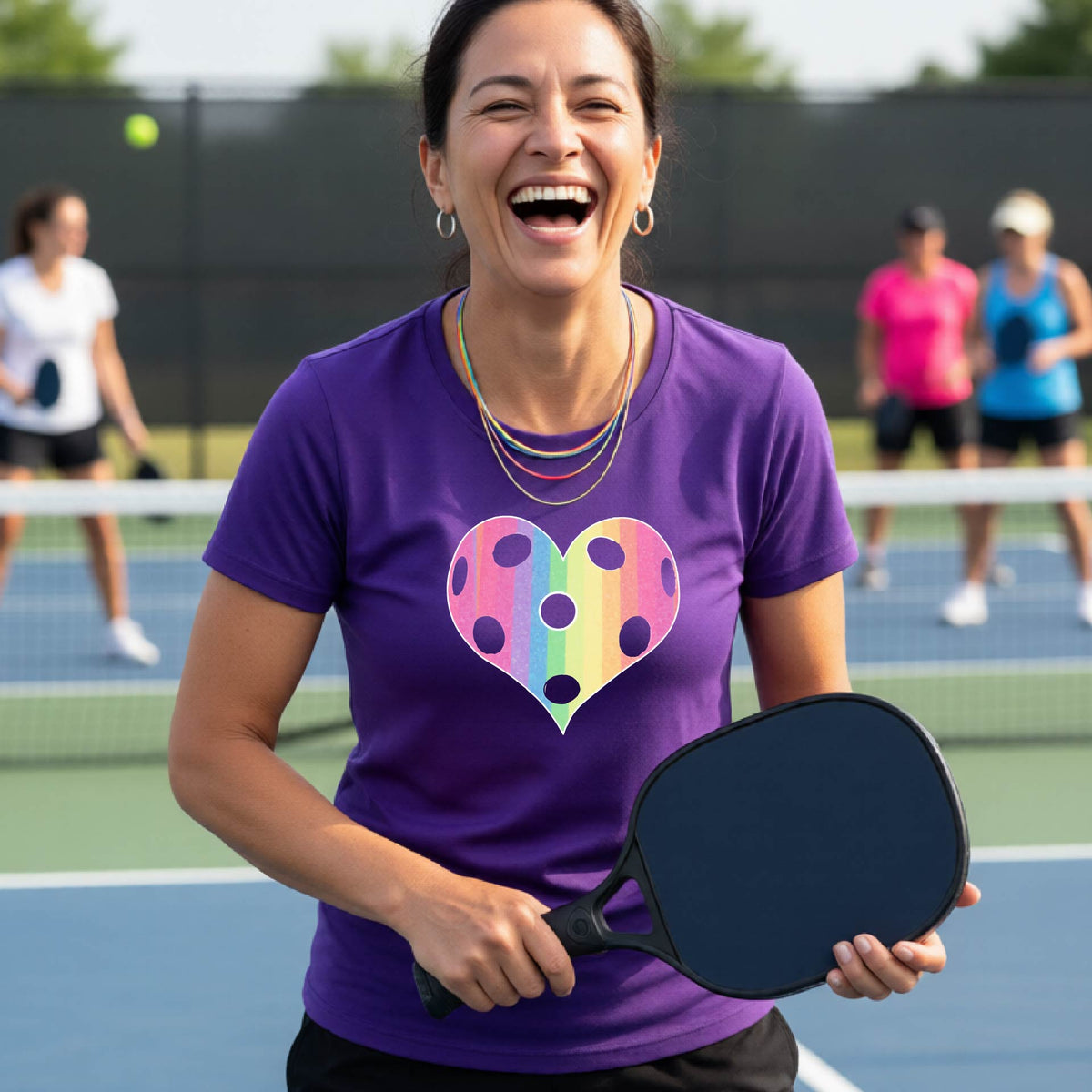 A woman laughing on a pickleball court, wearing a purple t-shirt with a graphic of a brightly rainbow-colored heart designed like a pickleball.