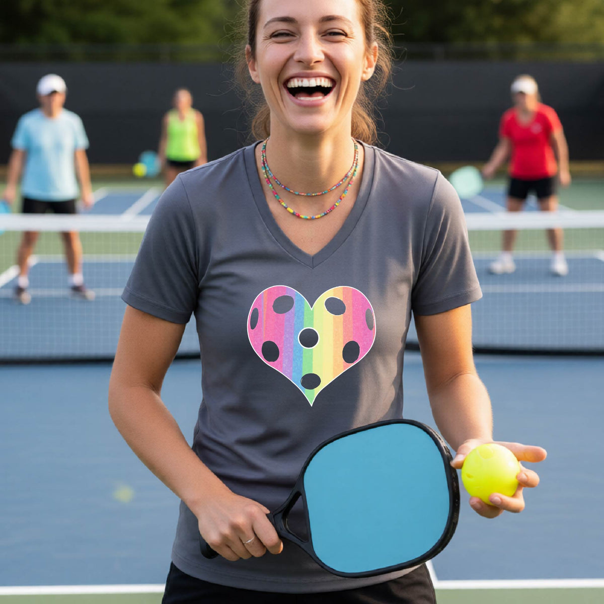 A woman laughing on a pickleball court, wearing a dark gray t-shirt with a graphic of a brightly rainbow-colored heart designed like a pickleball.