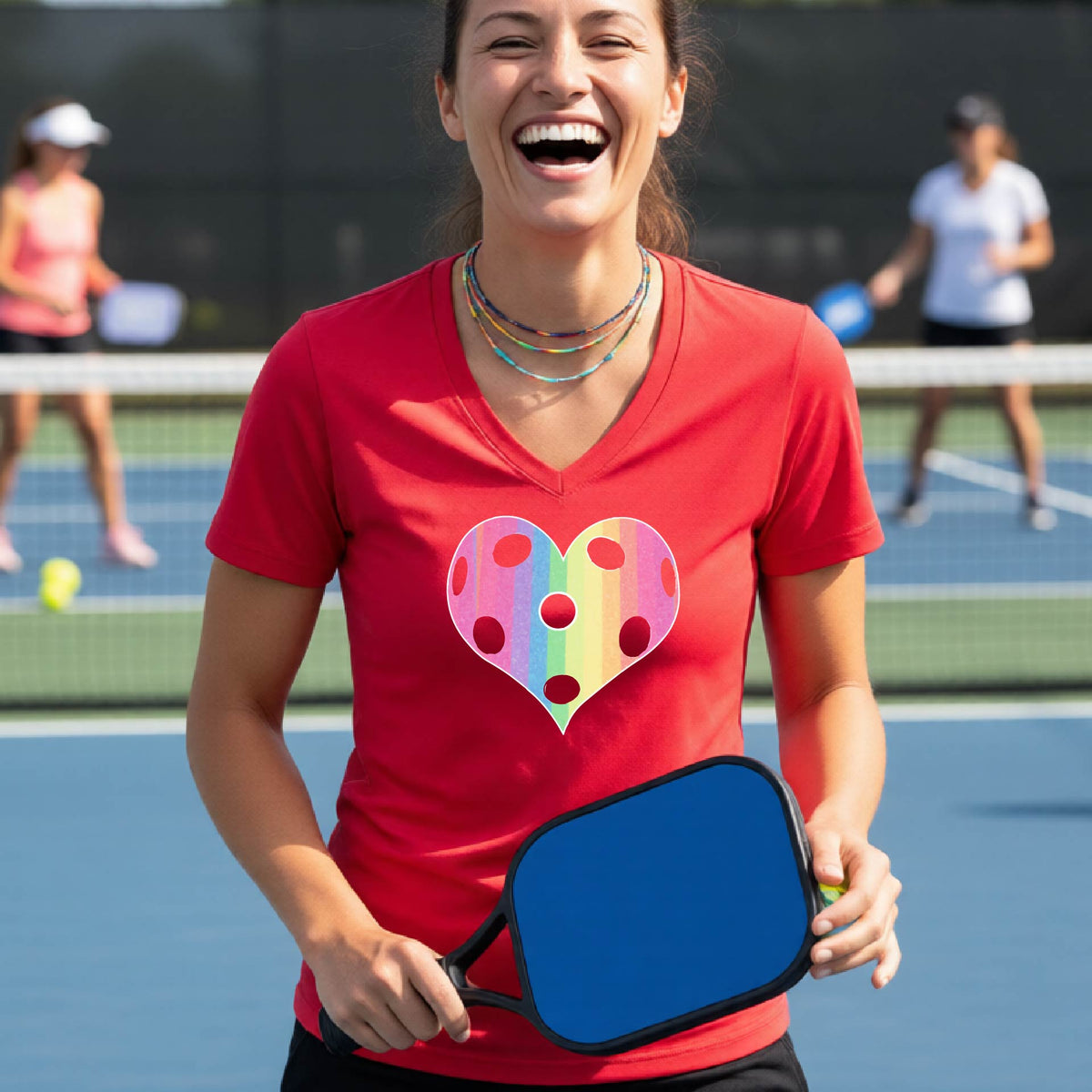 A woman laughing on a pickleball court, wearing a red t-shirt with a graphic of a brightly rainbow-colored heart designed like a pickleball.