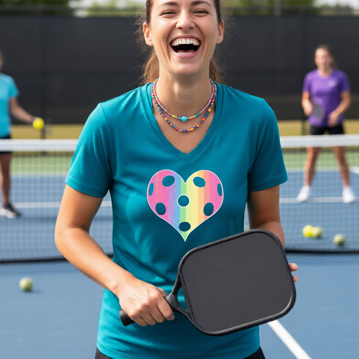 A woman laughing on a pickleball court, wearing a teal t-shirt with a graphic of a brightly rainbow-colored heart designed like a pickleball.