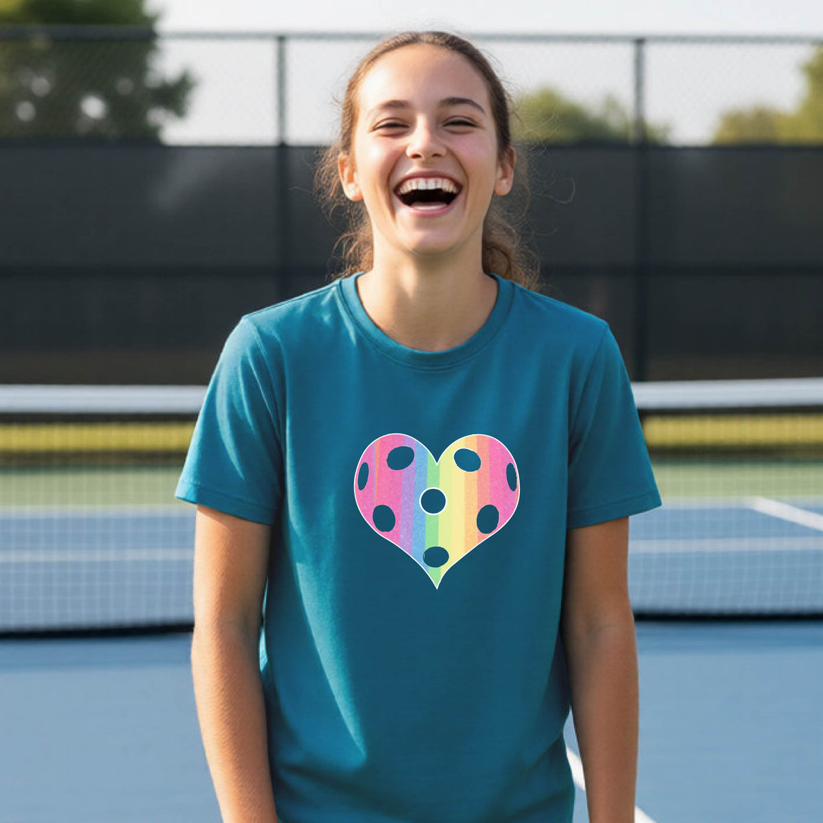 A young girl laughing on a pickleball court, wearing a teal t-shirt with a graphic of a brightly rainbow-colored heart designed like a pickleball.