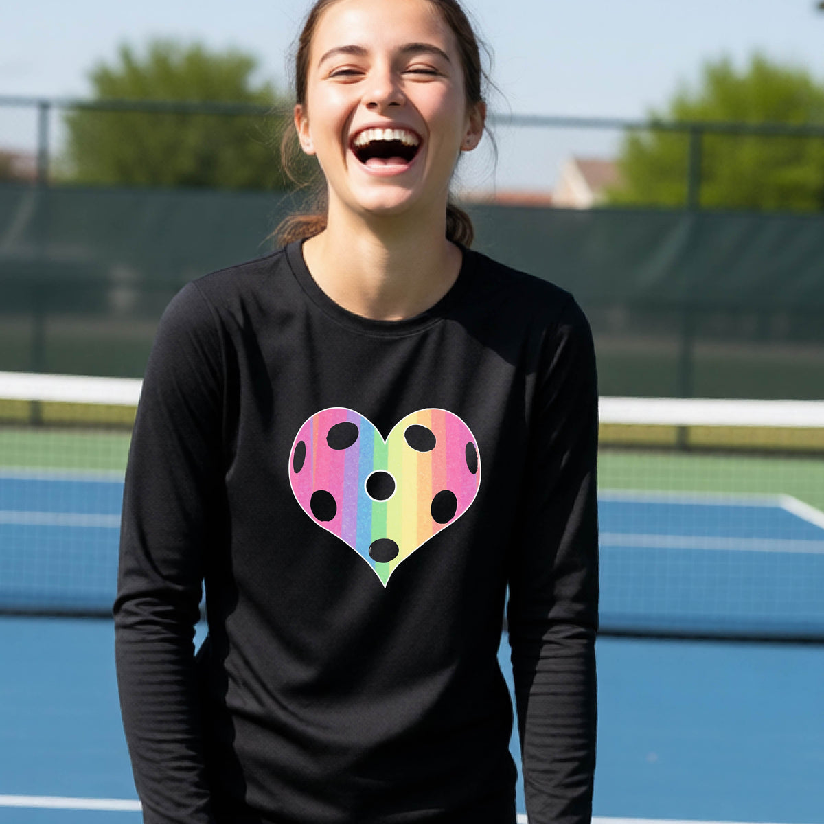 A young girl laughing on a pickleball court, wearing a black t-shirt with a graphic of a brightly rainbow-colored heart designed like a pickleball.
