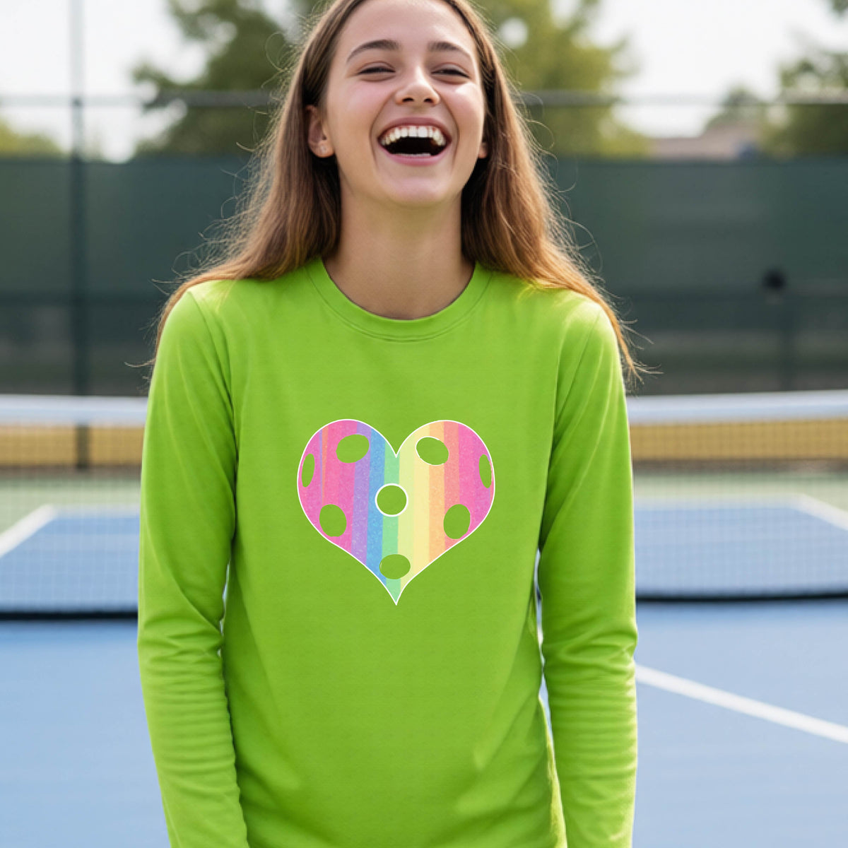 A young girl laughing on a pickleball court, wearing a lime green t-shirt with a graphic of a brightly rainbow-colored heart designed like a pickleball.