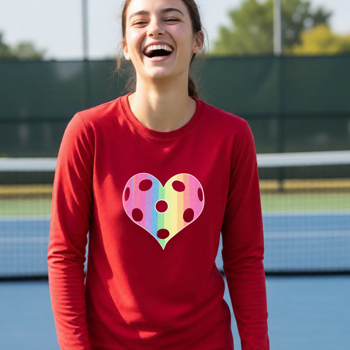 A young girl laughing on a pickleball court, wearing a red t-shirt with a graphic of a brightly rainbow-colored heart designed like a pickleball.