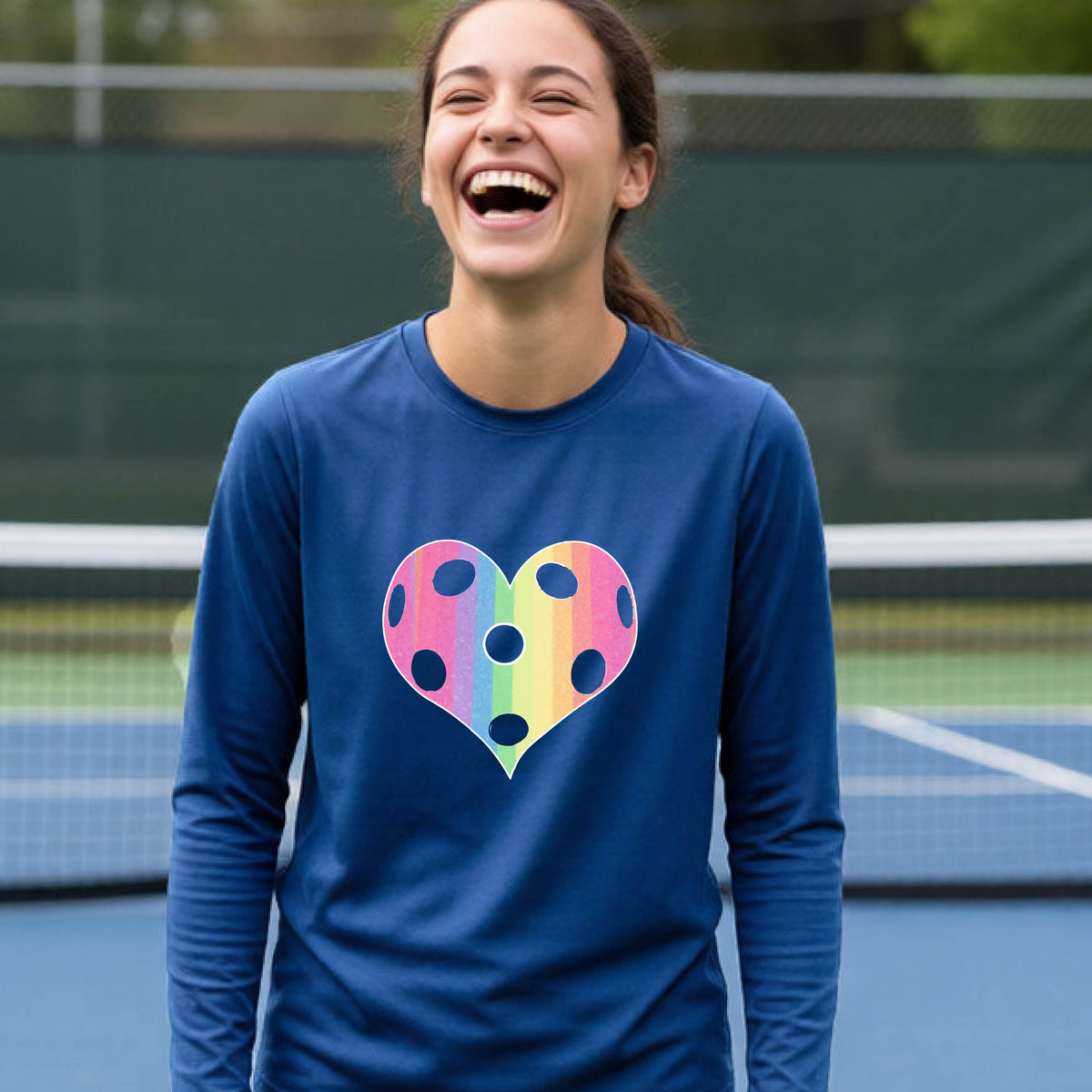 A young girl laughing on a pickleball court, wearing a royal blue t-shirt with a graphic of a brightly rainbow-colored heart designed like a pickleball.