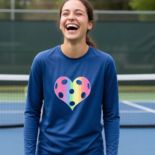 A young girl laughing on a pickleball court, wearing a royal blue t-shirt with a graphic of a brightly rainbow-colored heart designed like a pickleball.