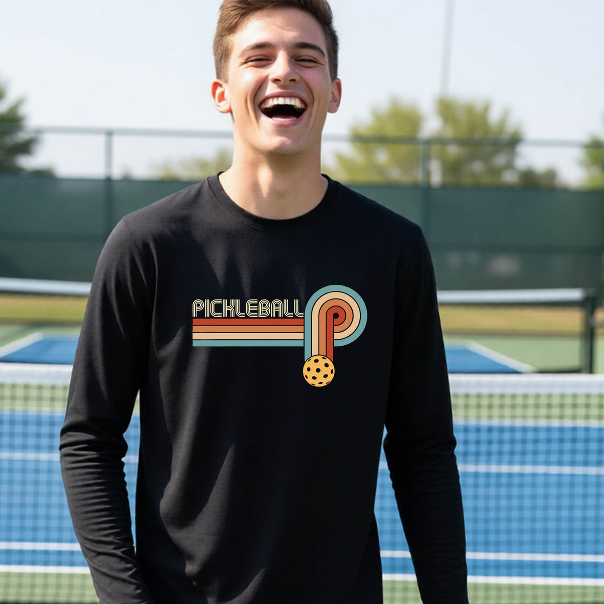 A young person on a pickleball court wearing a black short-sleeve t-shirt with a retro graphic of the word  “pickleball” on top of four colored lines that turn and go down ending in an orange pickleball, standing on a pickleball court.
