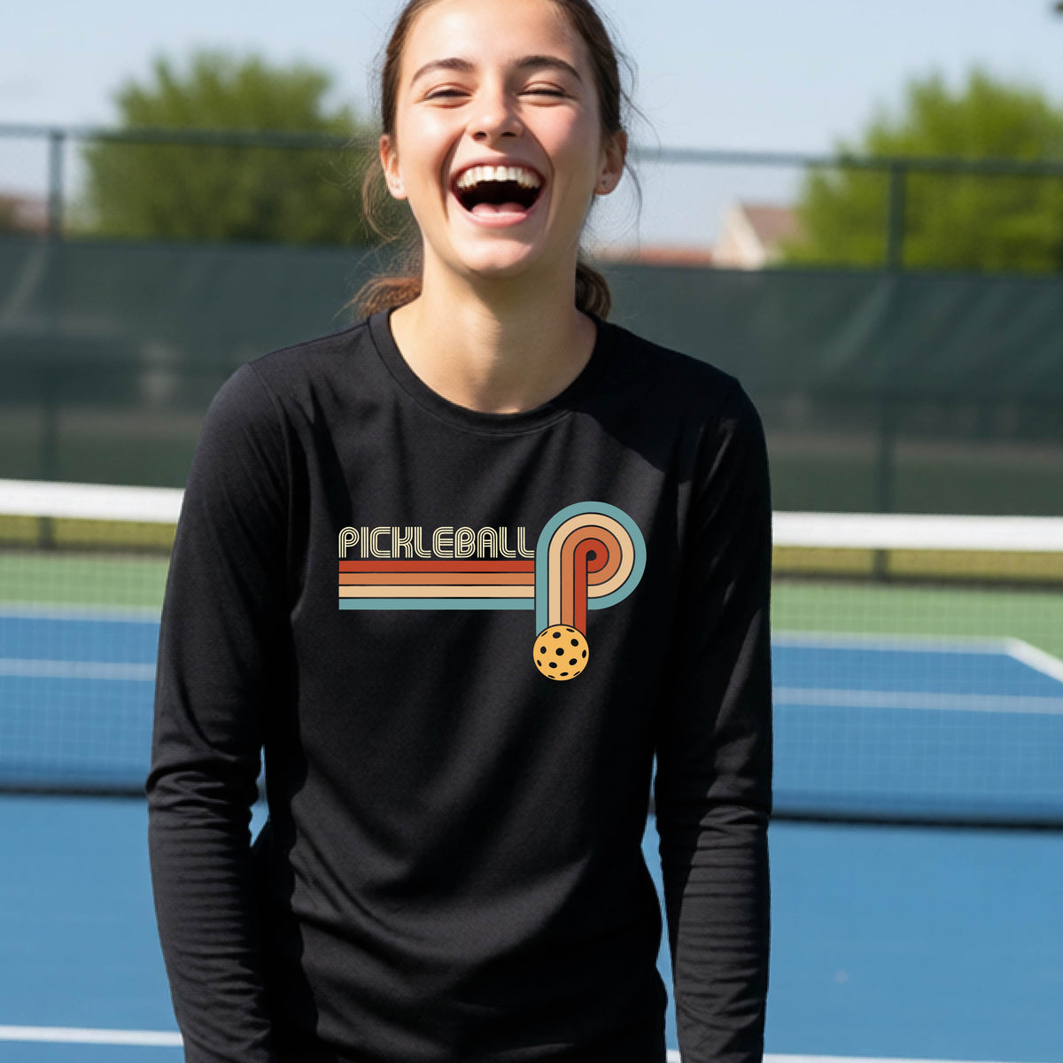 A young person on a pickleball court wearing a black short-sleeve t-shirt with a retro graphic of the word  “pickleball” on top of four colored lines that turn and go down ending in an orange pickleball, standing on a pickleball court.