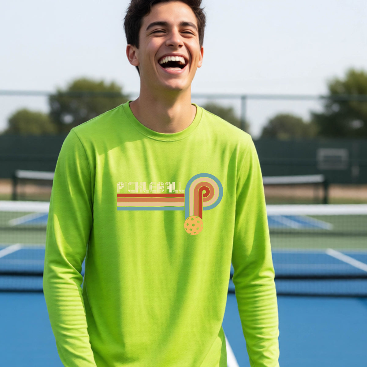 A young person on a pickleball court wearing a lime green short-sleeve t-shirt with a retro graphic of the word  “pickleball” on top of four colored lines that turn and go down ending in an orange pickleball, standing on a pickleball court.
