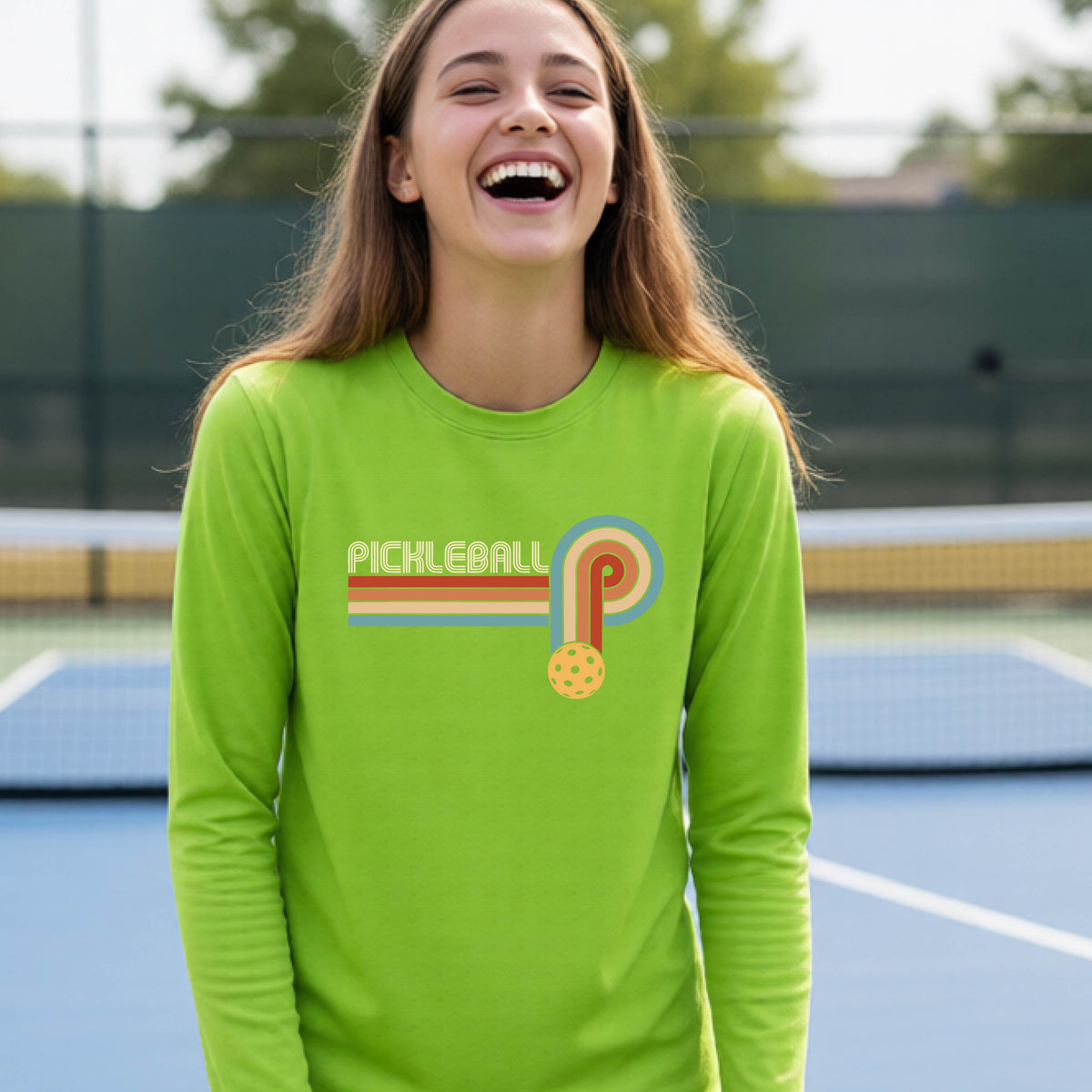 A young person on a pickleball court wearing a lime green short-sleeve t-shirt with a retro graphic of the word  “pickleball” on top of four colored lines that turn and go down ending in an orange pickleball, standing on a pickleball court.