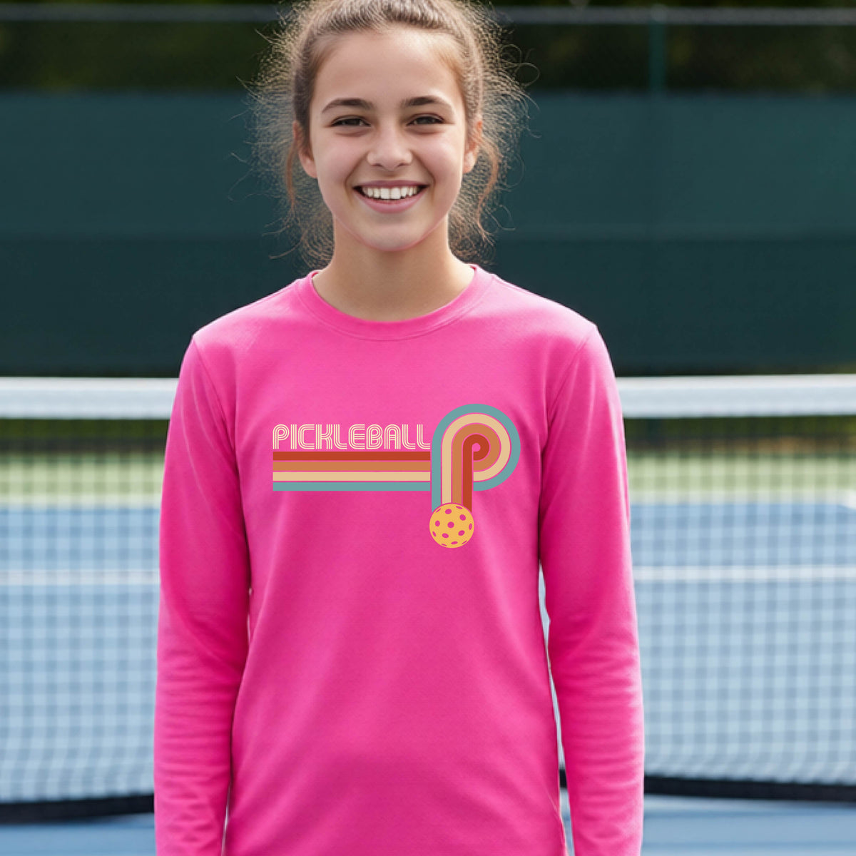 A young person on a pickleball court wearing a pink short-sleeve t-shirt with a retro graphic of the word  “pickleball” on top of four colored lines that turn and go down ending in an orange pickleball, standing on a pickleball court.