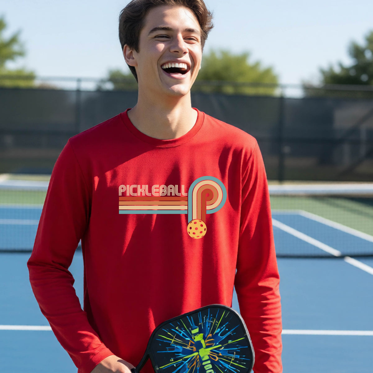 A young person on a pickleball court wearing a red short-sleeve t-shirt with a retro graphic of the word  “pickleball” on top of four colored lines that turn and go down ending in an orange pickleball, standing on a pickleball court.