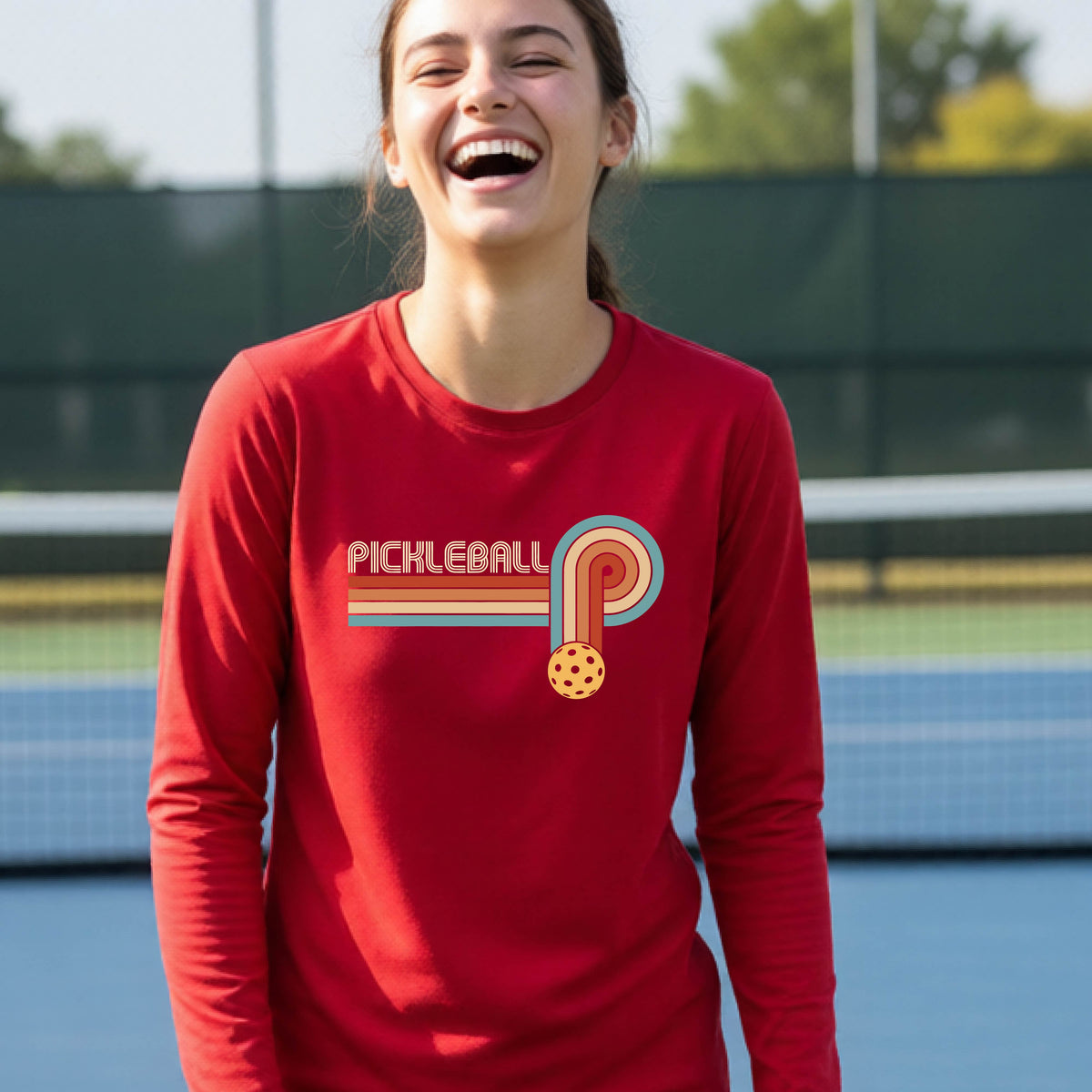 A young person on a pickleball court wearing a red short-sleeve t-shirt with a retro graphic of the word  “pickleball” on top of four colored lines that turn and go down ending in an orange pickleball, standing on a pickleball court.