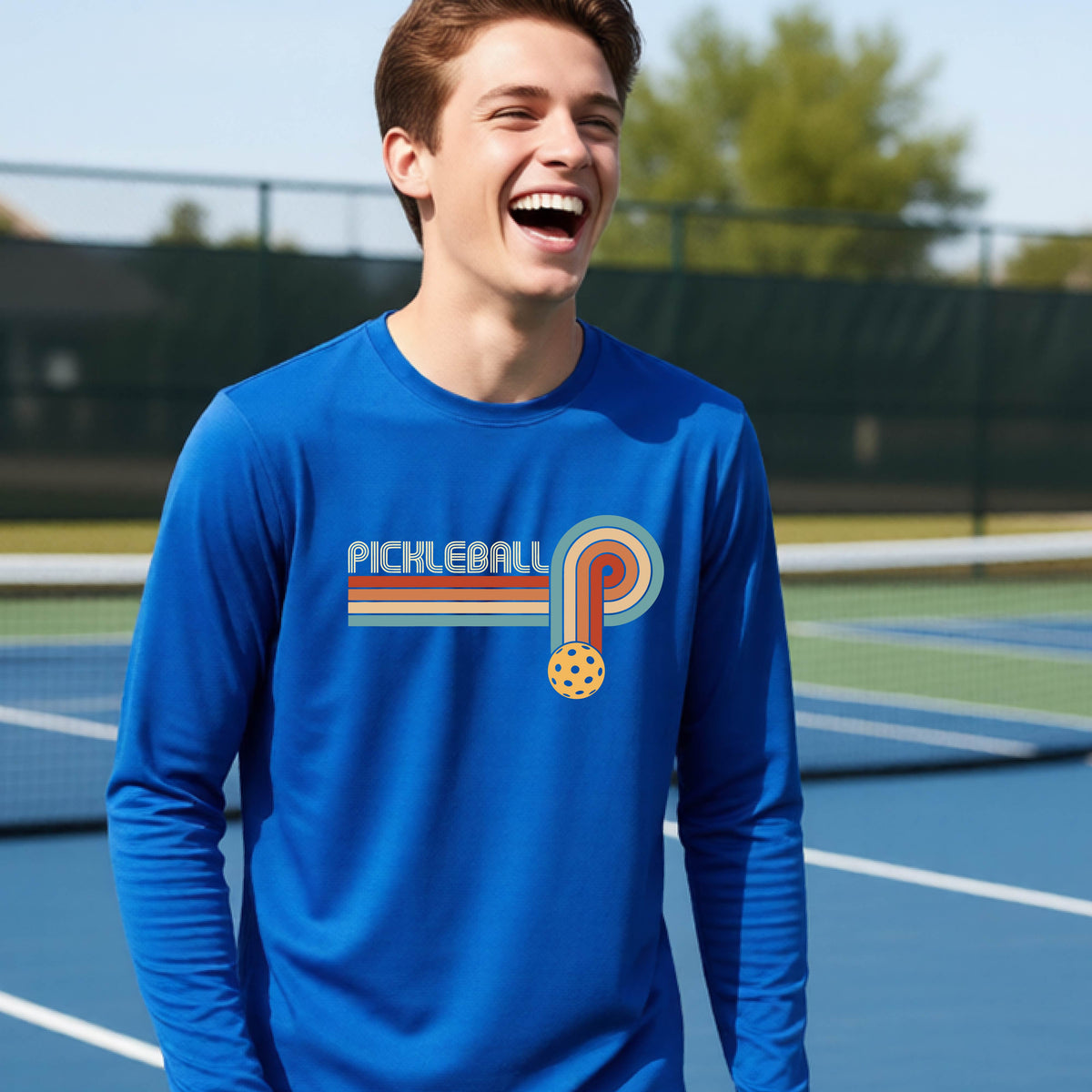 A young person on a pickleball court wearing a royal blue short-sleeve t-shirt with a retro graphic of the word  “pickleball” on top of four colored lines that turn and go down ending in an orange pickleball, standing on a pickleball court.