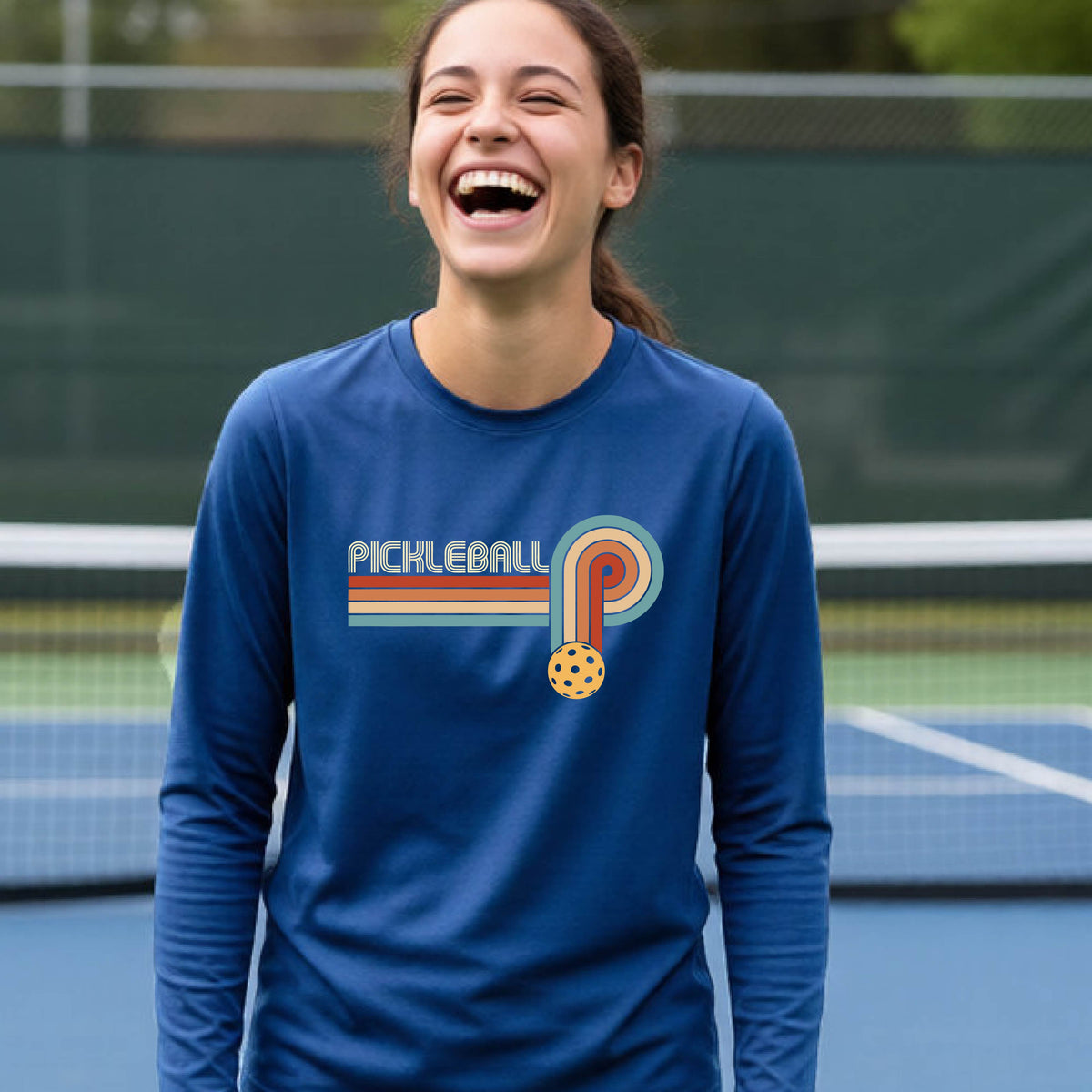 A young person on a pickleball court wearing a royal blue short-sleeve t-shirt with a retro graphic of the word  “pickleball” on top of four colored lines that turn and go down ending in an orange pickleball, standing on a pickleball court.