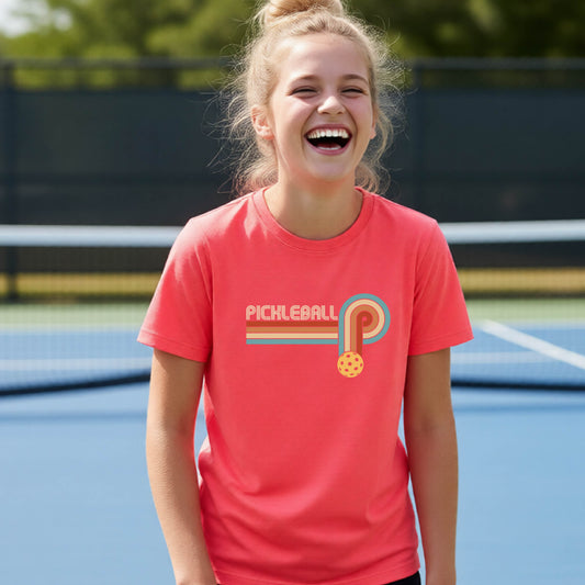 A young person on a pickleball court wearing a coral pink short-sleeve t-shirt with a retro graphic of the word  “pickleball” on top of four colored lines that turn and go down ending in an orange pickleball, standing on a pickleball court.