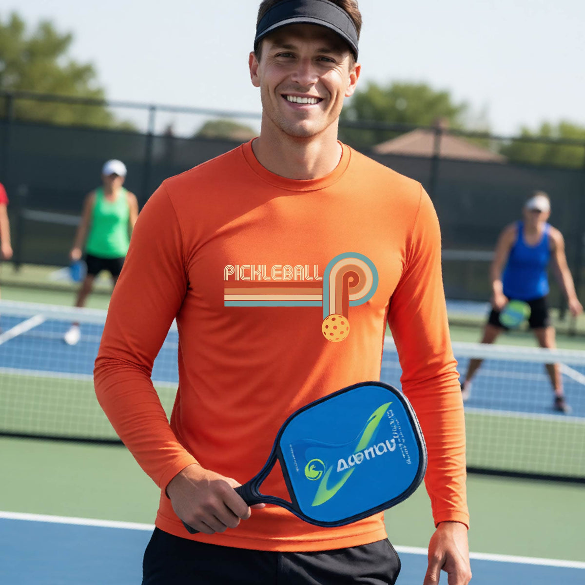 A man on a pickleball court wearing a bright orange long-sleeve t-shirt with a retro graphic of the word  “pickleball” on top of four colored lines that turn and go down ending in an orange pickleball, standing on a pickleball court.
