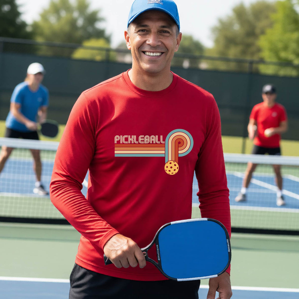 A man on a pickleball court wearing a red long-sleeve t-shirt with a retro graphic of the word  “pickleball” on top of four colored lines that turn and go down ending in an orange pickleball, standing on a pickleball court.