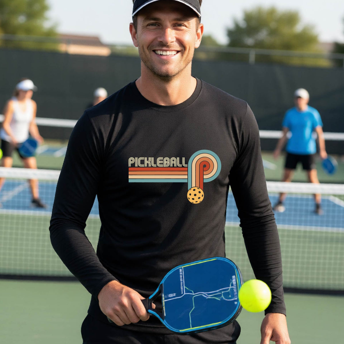A man on a pickleball court wearing a black long-sleeve t-shirt with a retro graphic of the word  “pickleball” on top of four colored lines that turn and go down ending in an orange pickleball, standing on a pickleball court.