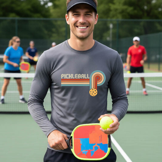 A man on a pickleball court wearing a gray long-sleeve t-shirt with a retro graphic of the word  “pickleball” on top of four colored lines that turn and go down ending in an orange pickleball, standing on a pickleball court.