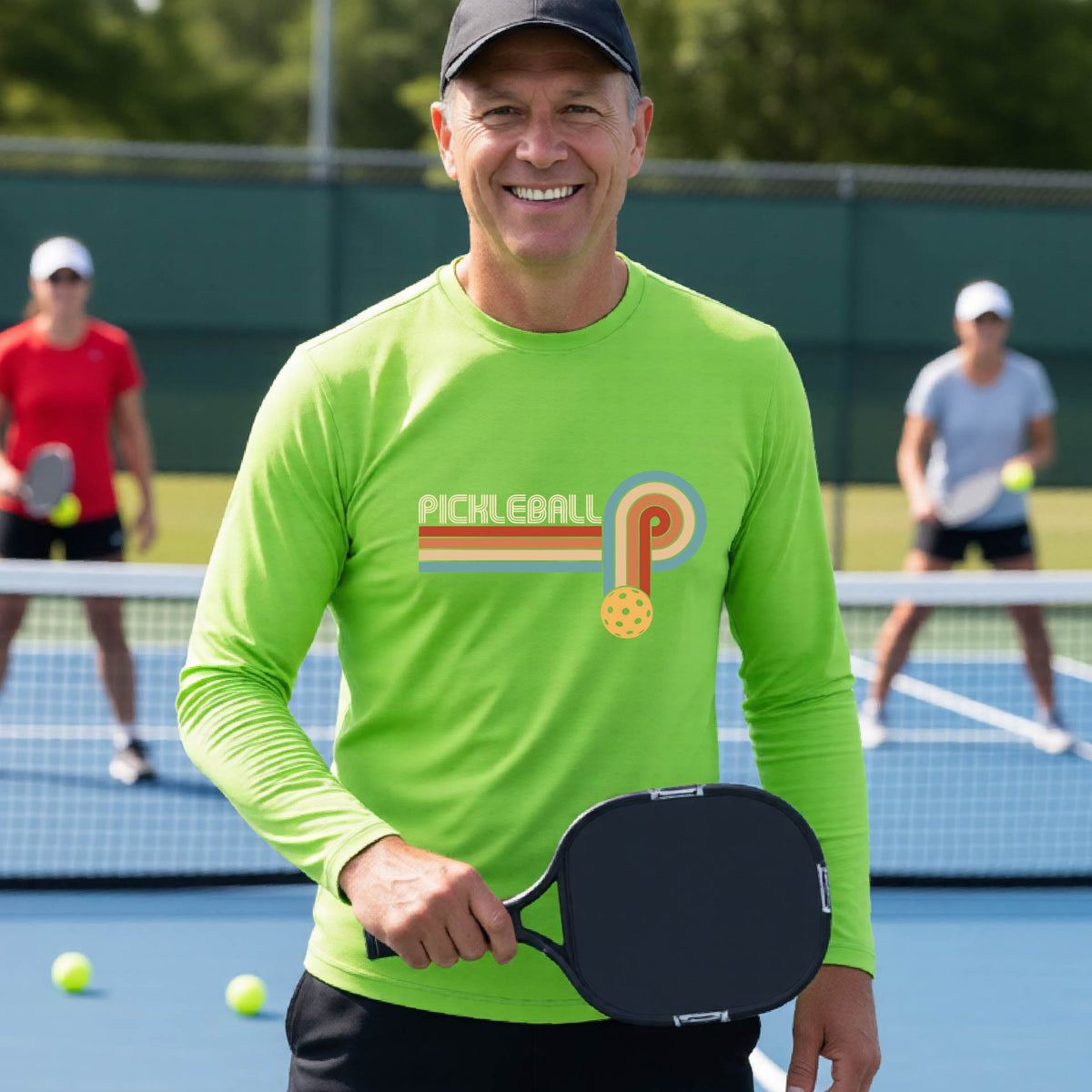 A man on a pickleball court wearing a lime green long-sleeve t-shirt with a retro graphic of the word  “pickleball” on top of four colored lines that turn and go down ending in an orange pickleball, standing on a pickleball court.