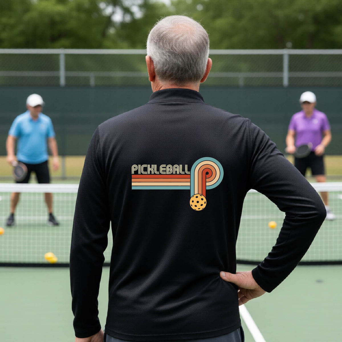 A man on a pickleball court showing the back of a black quarter-zip shirt with a retro graphic of the word  “pickleball” on top of four colored lines that turn and go down ending in an orange pickleball, standing on a pickleball court.