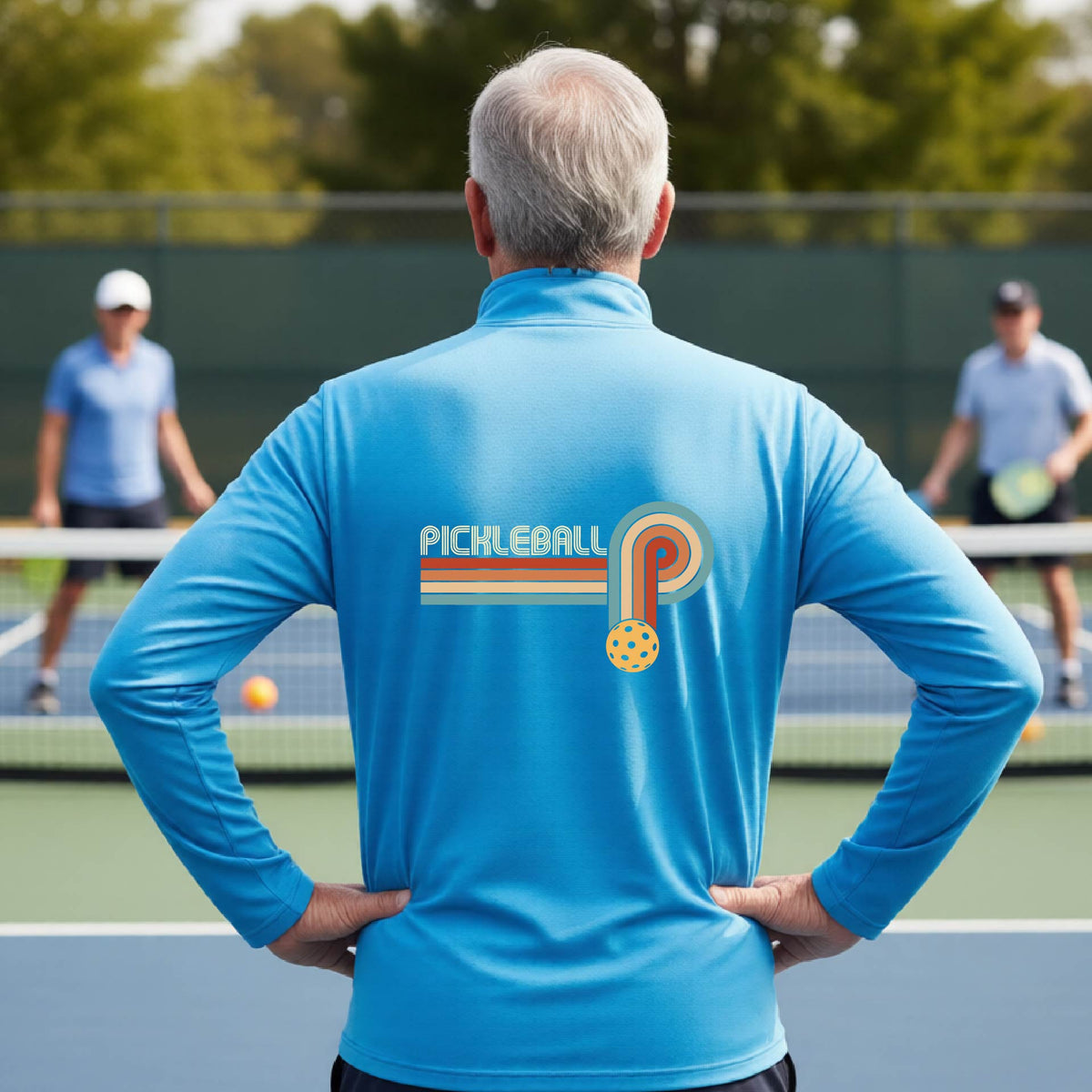 A man on a pickleball court showing the back of a bright blue quarter-zip shirt with a retro graphic of the word  “pickleball” on top of four colored lines that turn and go down ending in an orange pickleball, standing on a pickleball court.
