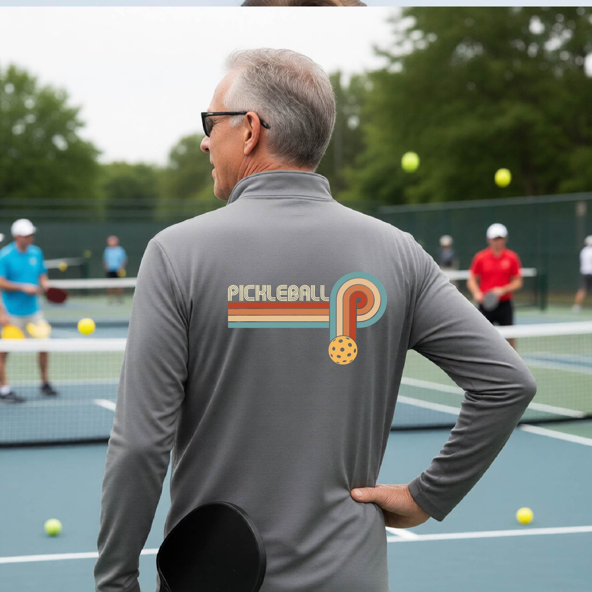 A man on a pickleball court showing the back of a gray quarter-zip shirt with a retro graphic of the word  “pickleball” on top of four colored lines that turn and go down ending in an orange pickleball, standing on a pickleball court.