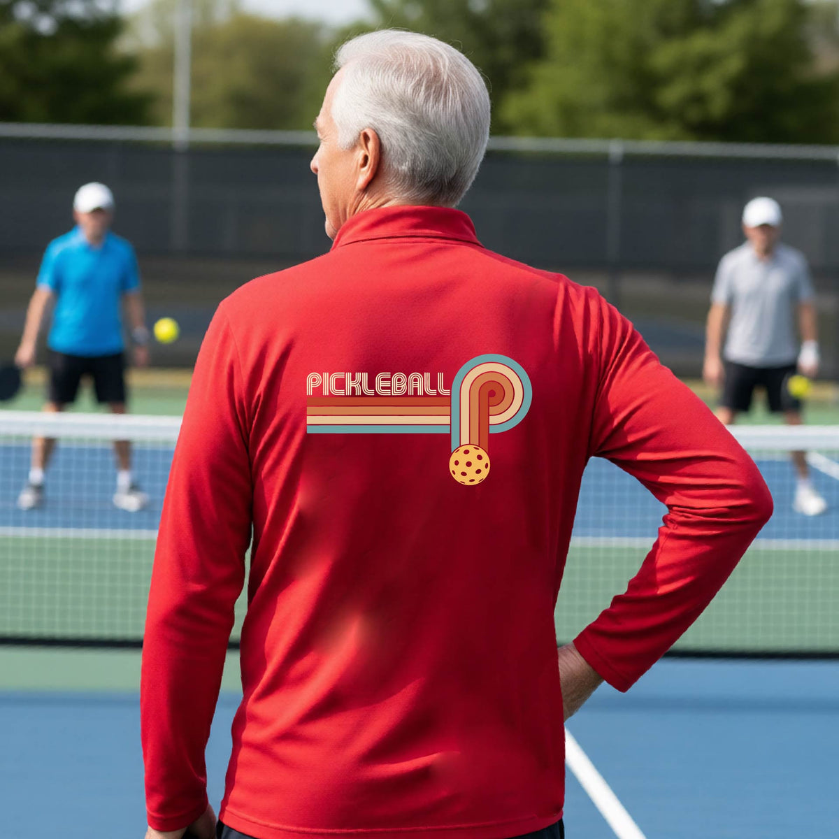 A man on a pickleball court showing the back of a red quarter-zip shirt with a retro graphic of the word  “pickleball” on top of four colored lines that turn and go down ending in an orange pickleball, standing on a pickleball court.