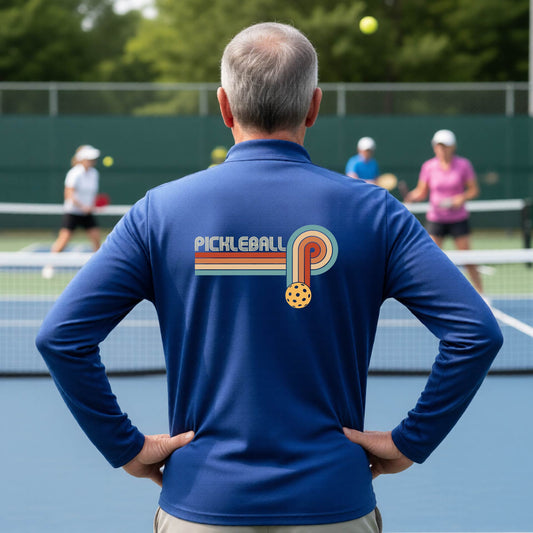 A man on a pickleball court showing the back of a royal blue quarter-zip shirt with a retro graphic of the word  “pickleball” on top of four colored lines that turn and go down ending in an orange pickleball, standing on a pickleball court.