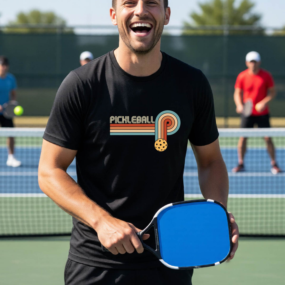 A man on a pickleball court wearing a black short-sleeve t-shirt with a retro graphic of the word  “pickleball” on top of four colored lines that turn and go down ending in an orange pickleball, standing on a pickleball court.