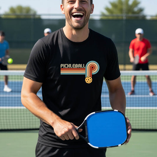 A man on a pickleball court wearing a black short-sleeve t-shirt with a retro graphic of the word  “pickleball” on top of four colored lines that turn and go down ending in an orange pickleball, standing on a pickleball court.