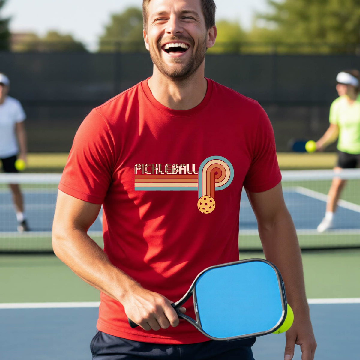 A man on a pickleball court wearing a red short-sleeve t-shirt with a retro graphic of the word  “pickleball” on top of four colored lines that turn and go down ending in an orange pickleball, standing on a pickleball court.