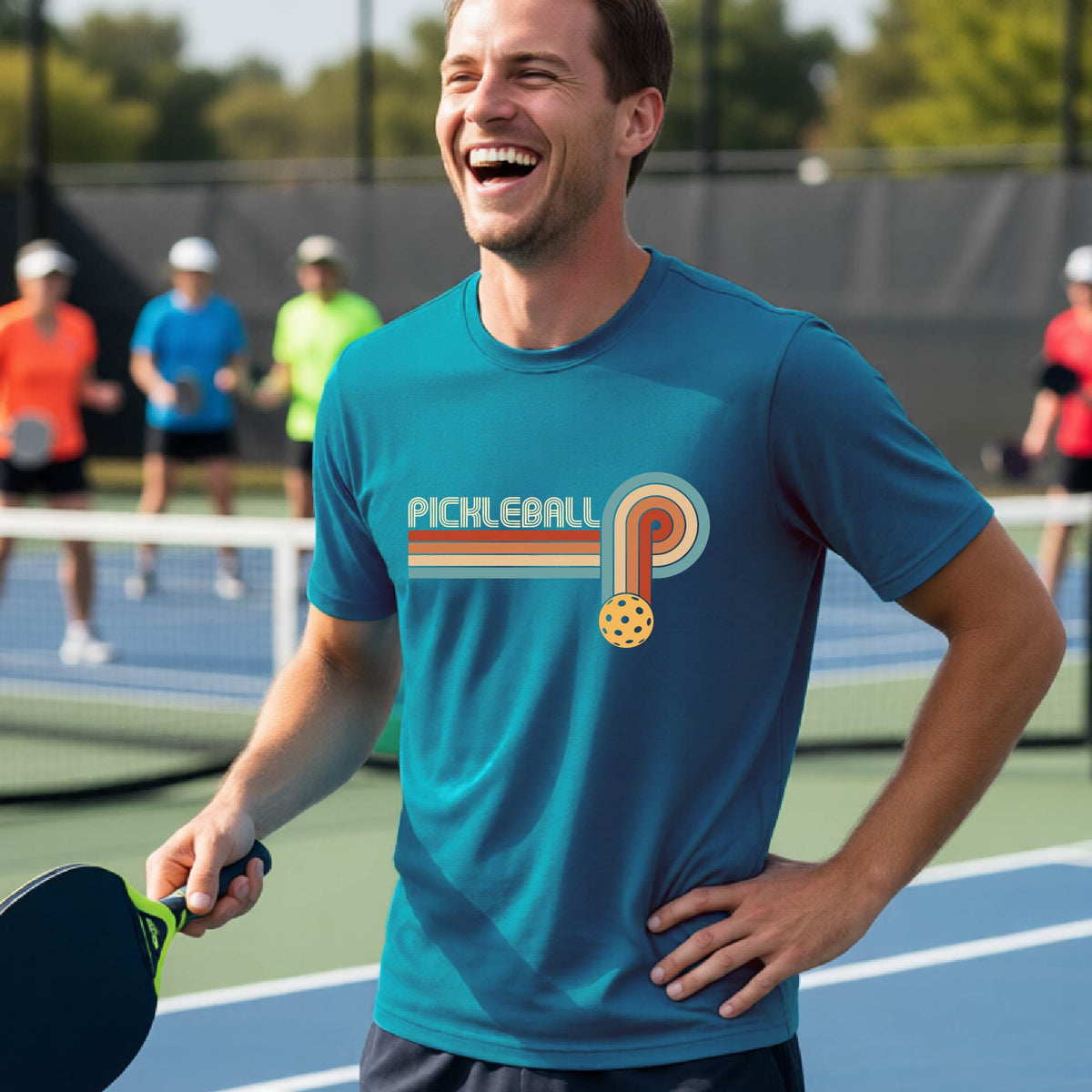 A man on a pickleball court wearing a teal short-sleeve t-shirt with a retro graphic of the word  “pickleball” on top of four colored lines that turn and go down ending in an orange pickleball, standing on a pickleball court.