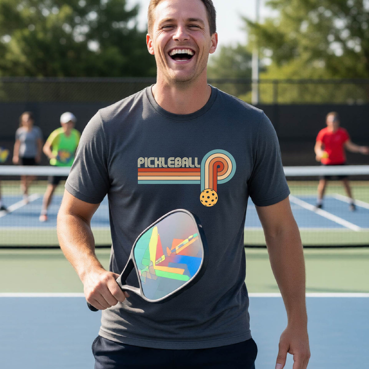 A man on a pickleball court wearing a gray short-sleeve t-shirt with a retro graphic of the word  “pickleball” on top of four colored lines that turn and go down ending in an orange pickleball, standing on a pickleball court.