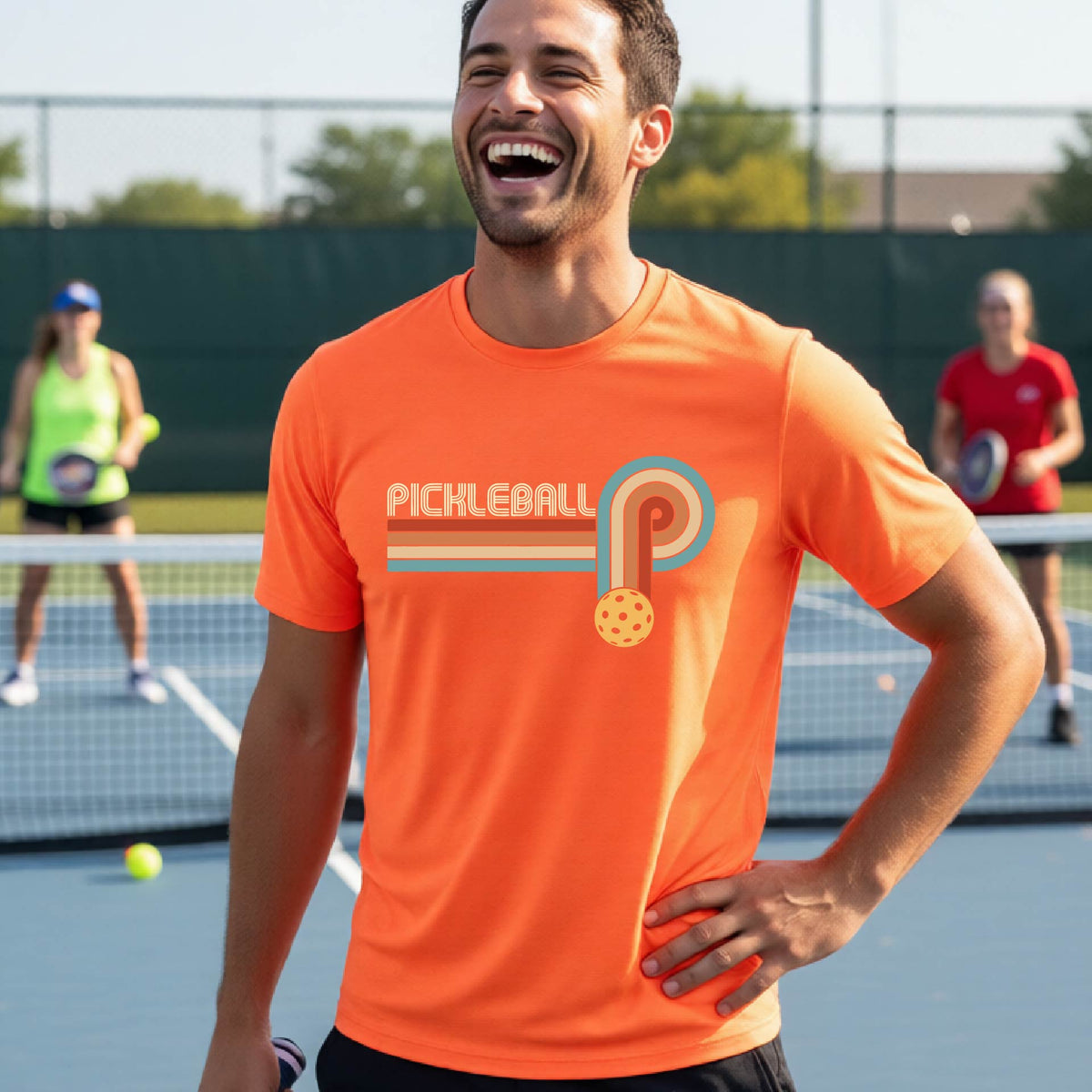 A man on a pickleball court wearing an orange short-sleeve t-shirt with a retro graphic of the word  “pickleball” on top of four colored lines that turn and go down ending in an orange pickleball, standing on a pickleball court.